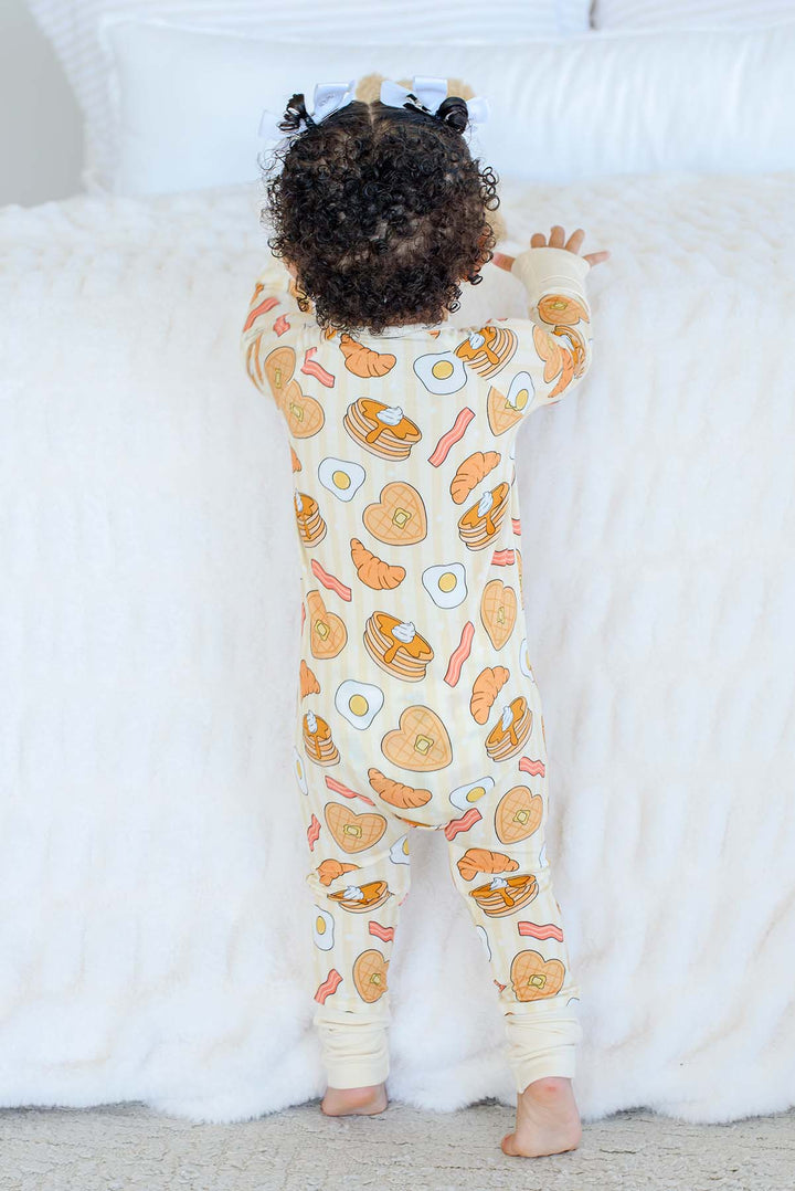 Back view of toddler standing in Caden Lane zip footie pajama with breakfast-themed print, hair in white bows