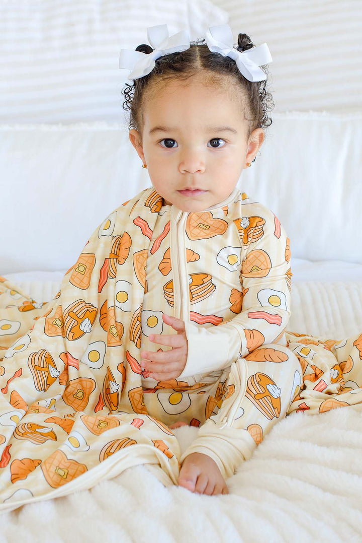Toddler girl sitting on a bed in Caden Lane zip footie pajama with matching blanket, pancake, egg, bacon, waffle and croissant print on cream stripes food print 