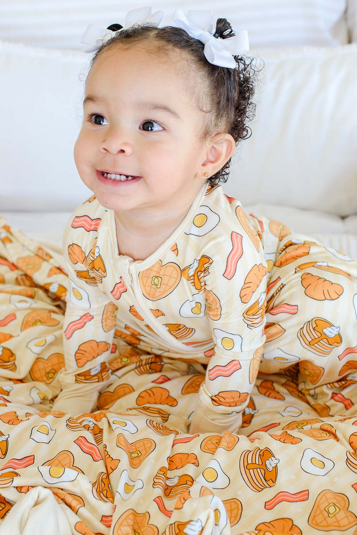 A baby girl wearing a Caden Lane cream-colored long-sleeve pajama set with a breakfast food print (eggs, bacon, pancakes) and a white bow in her hair, sitting on a white blanket.