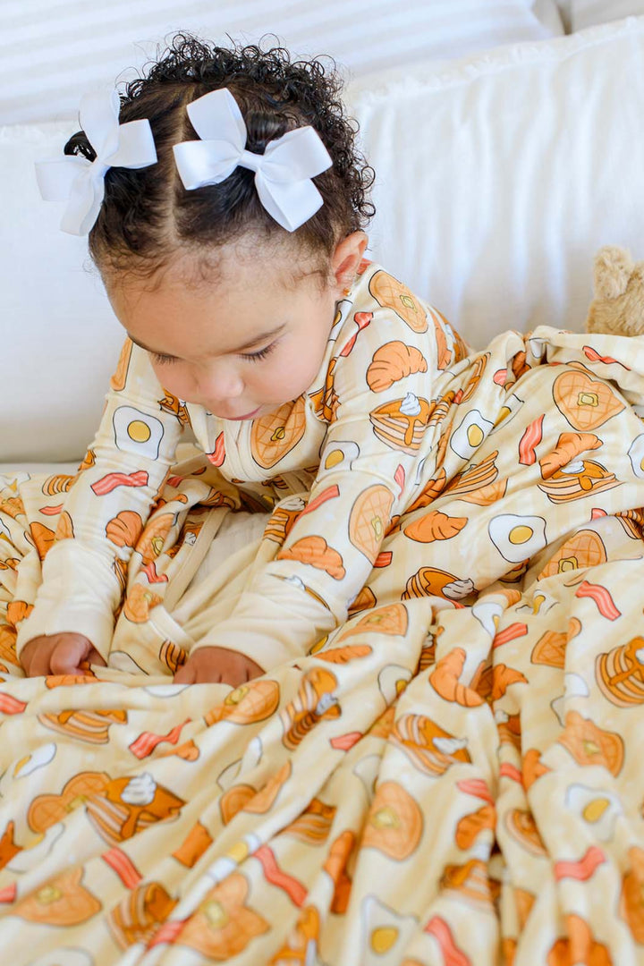 A toddler girl with curly hair and white bow headbands is lying on a white bed, wrapped in a cream-colored blanket and wearing a matching long-sleeve top printed with a breakfast food pattern of eggs, bacon, pancakes, and toast.
