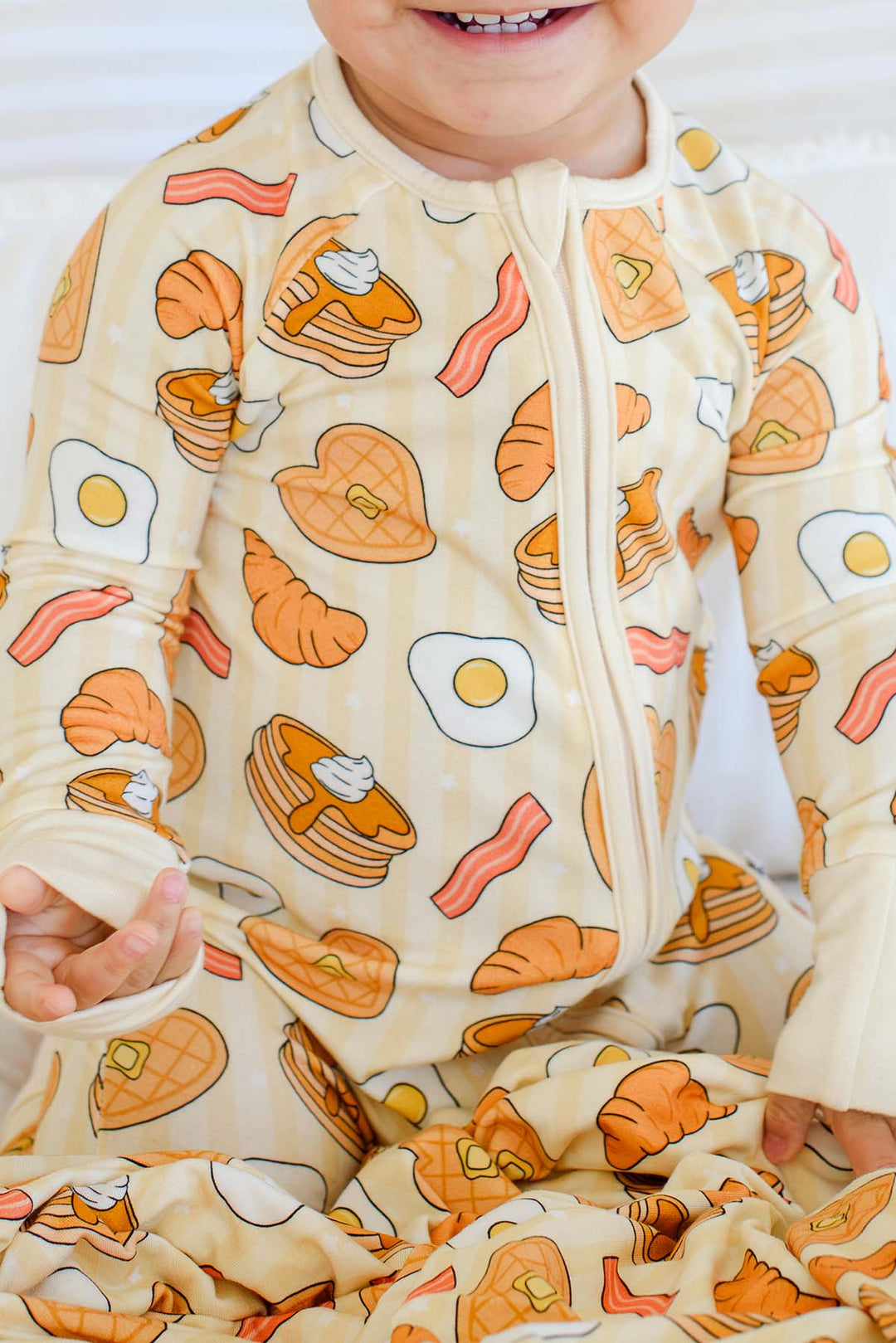 Close-up of smiling toddler in Caden Lane zip footie pajama, showing pancake, bacon and croissant print detail