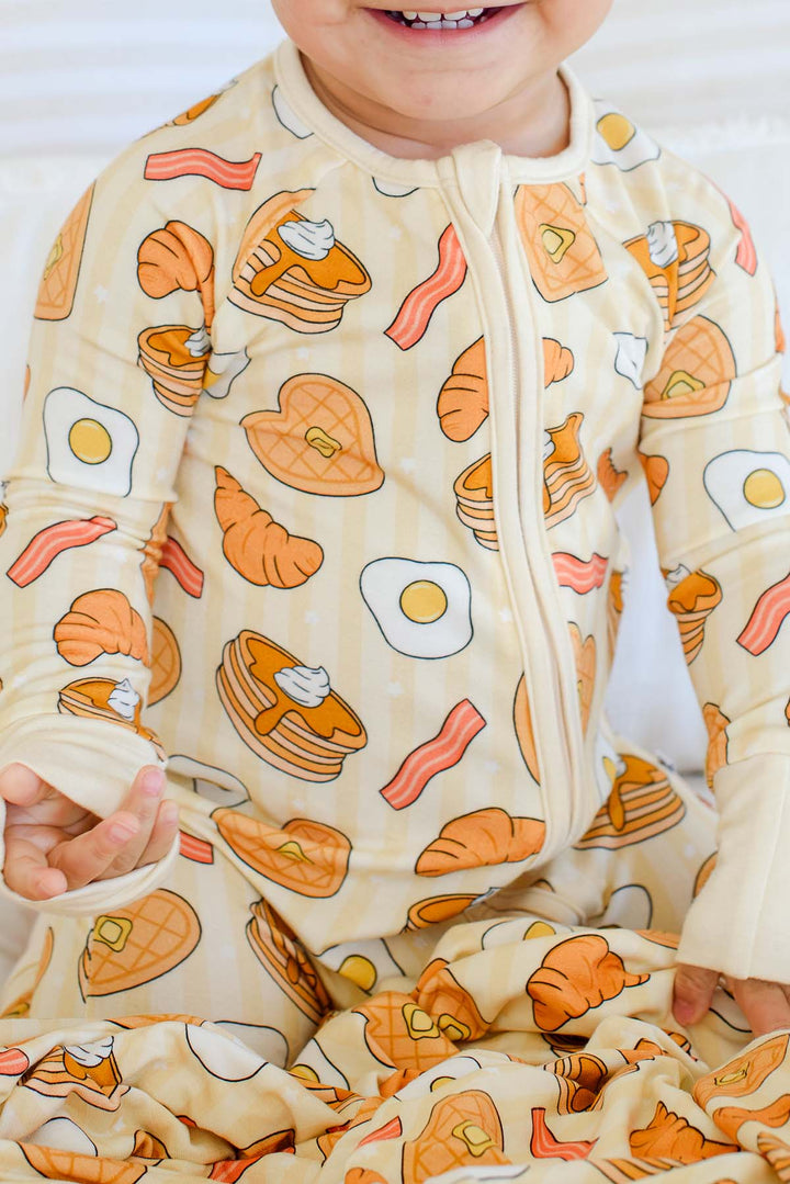 Close-up of smiling toddler in Caden Lane zip footie pajama, showing pancake, bacon and croissant print detail