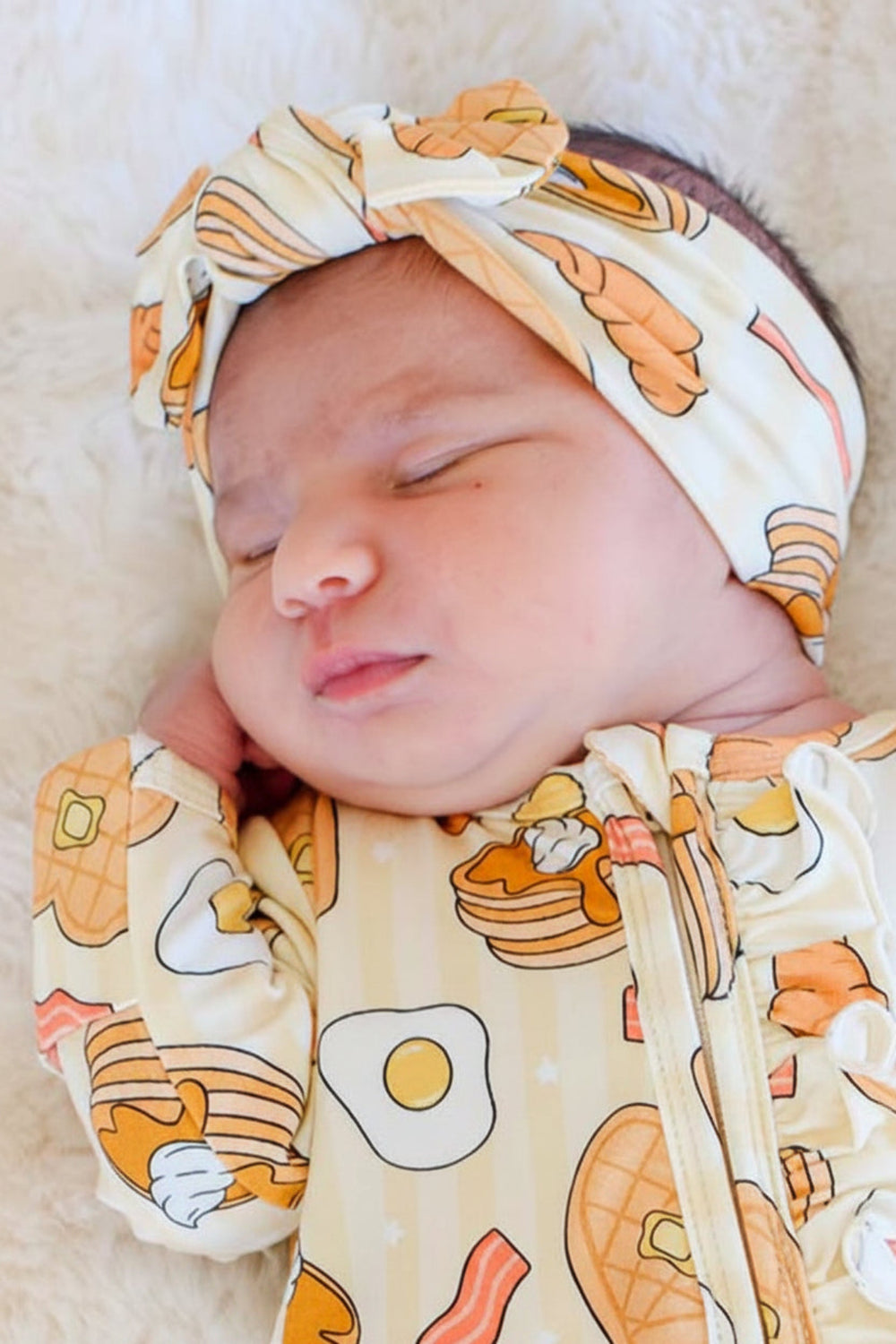 A sleeping newborn baby wearing a Caden Lane cream-colored footed sleeper and matching headband with a breakfast food print of pancakes, bacon, eggs, and toast, lying on a white faux fur blanket.