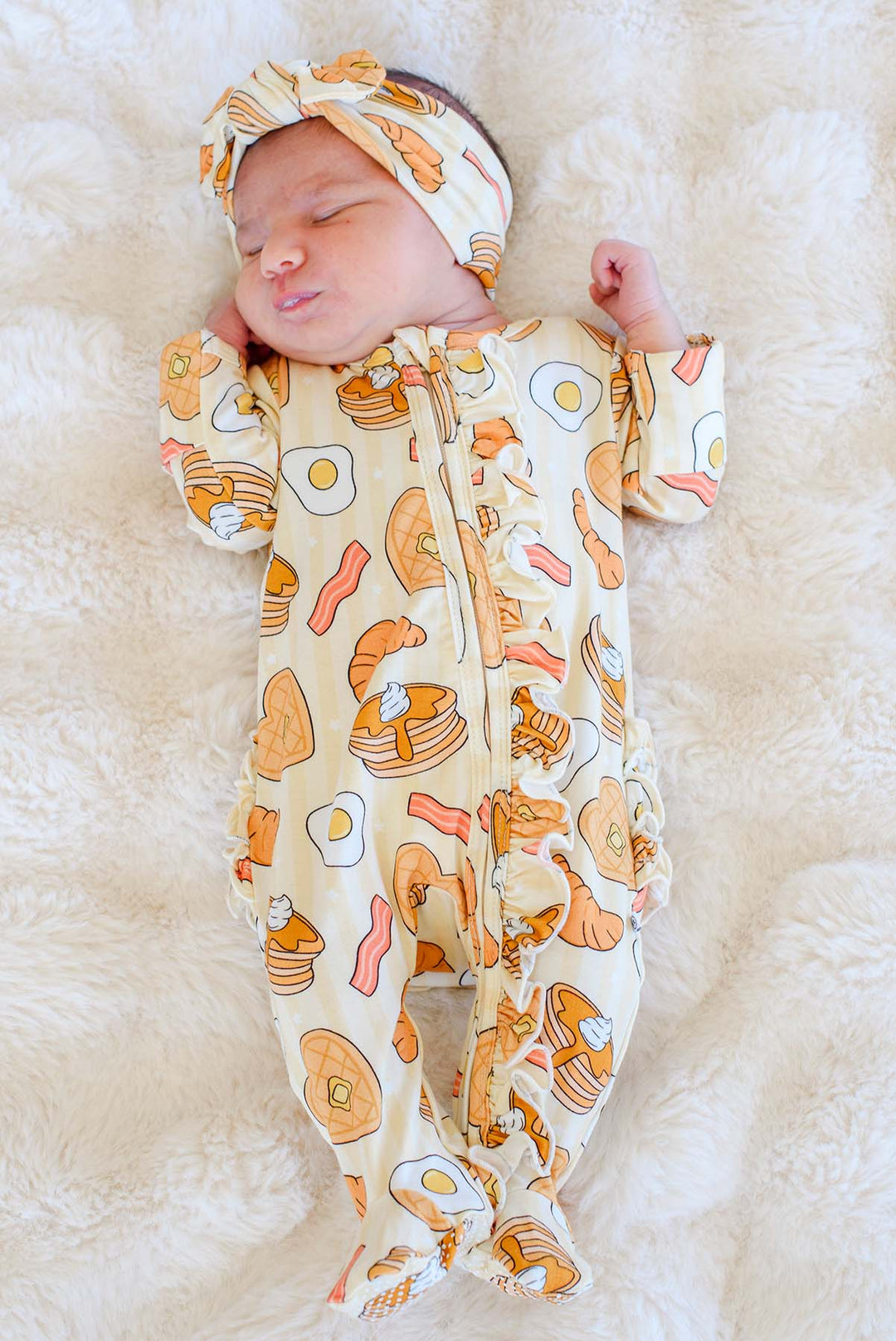 Newborn wearing Caden Lane I Love You A Brunch ruffle footie and matching knotted headband, sleeping on faux fur