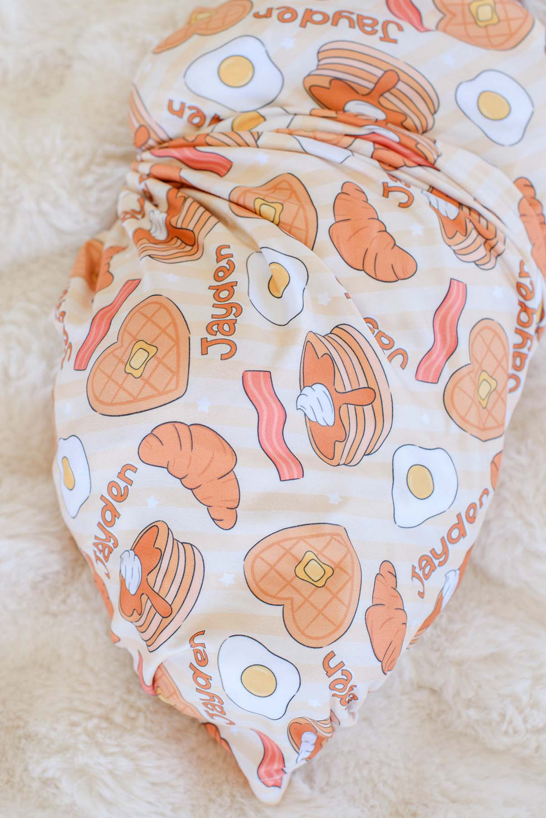 Close-up of Caden Lane personalized swaddle blanket, showing "Jayden" name repeated among pancake, egg, bacon and croissant print