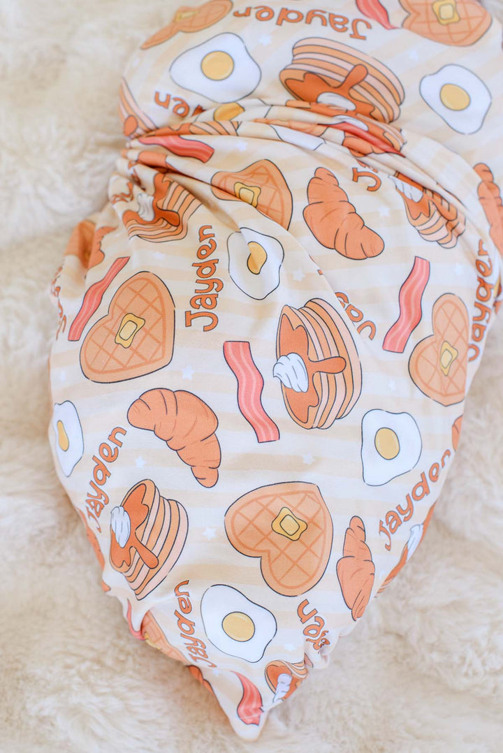 Close-up of Caden Lane personalized swaddle blanket, showing "Jayden" name repeated among pancake, egg, bacon and croissant print