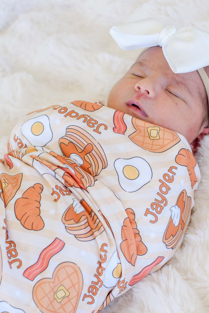 Close-up of sleeping newborn in white bow headband, wrapped in Caden Lane personalized "Jayden" breakfast-print swaddle