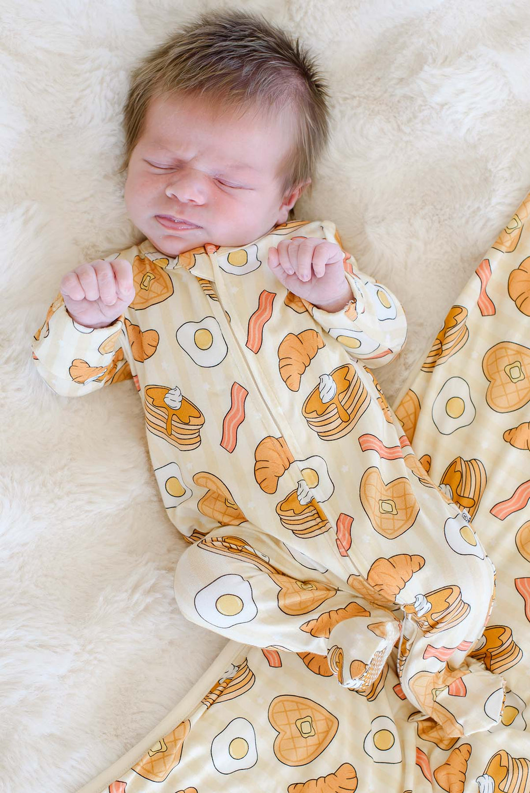 Newborn sleeping while wearing Caden Lane's "Love You A Brunch" print zip-up pajamas, featuring waffles, eggs, bacon, and croissants.