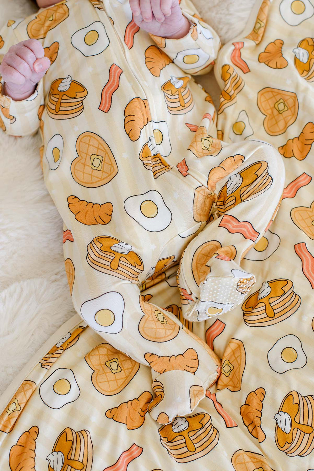 Close-up of newborn wearing Caden Lane's "Love You A Brunch" print zip-up pajamas, featuring waffles, eggs, bacon, and croissants.