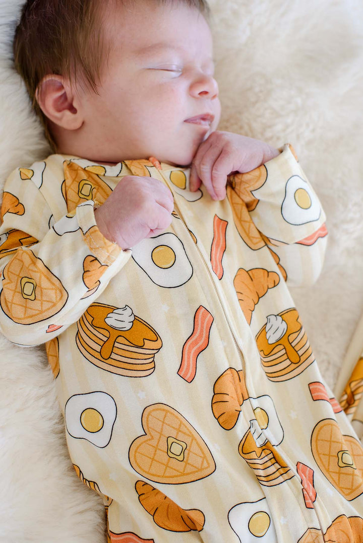 Close-up of newborn sleeping while wearing Caden Lane's "I Love You Brunch" print zip-up pajamas, featuring waffles, eggs, bacon, and croissants.