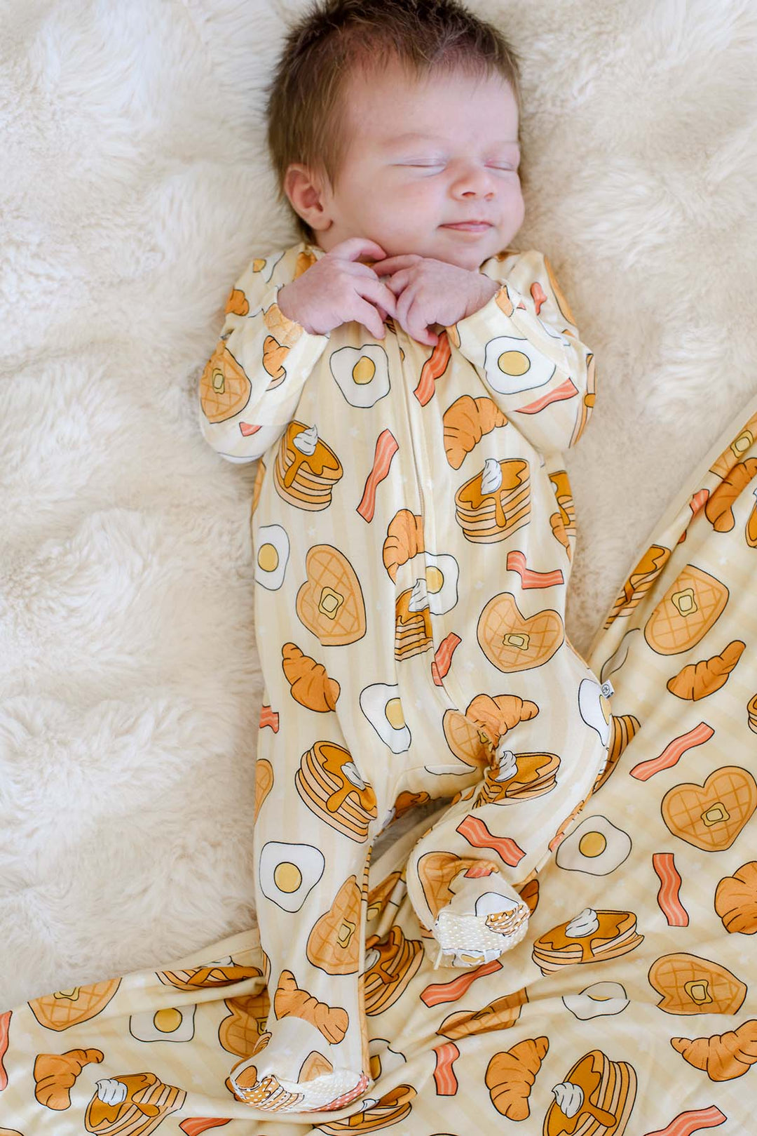 Newborn sleeping while wearing Caden Lane's "I Love You Brunch" print zip-up pajamas, featuring waffles, eggs, bacon, and croissants.
