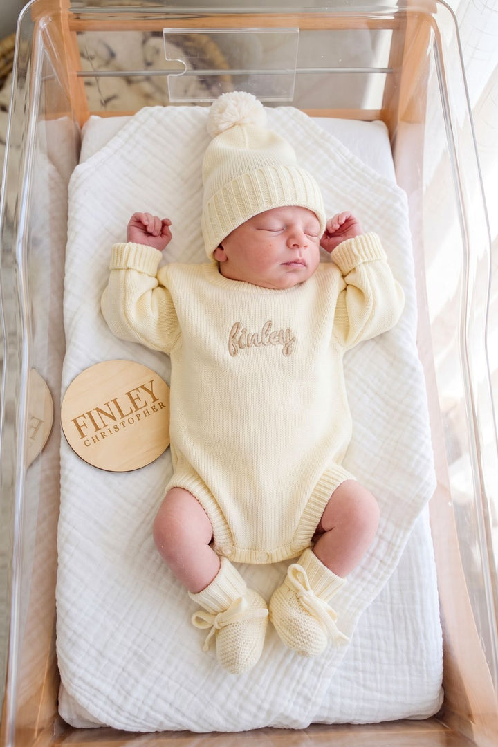 A sleeping newborn baby wearing a cream knit onesie with 'Finley' embroidered on the chest, a matching knit hat, and booties, lying on a white muslin blanket inside a clear baby bassinet. A round wooden tag reading 'FINLEY CHRISTOPHER' rests on the blanket.