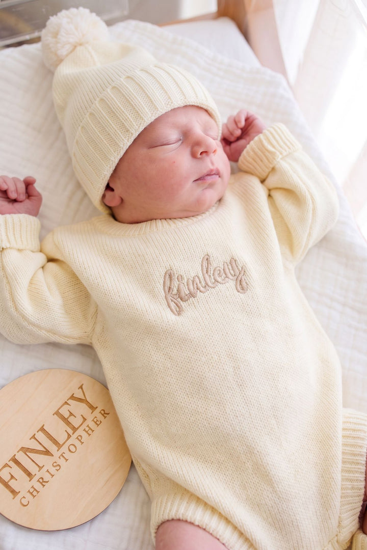 A sleeping newborn baby wearing a Caden Lane cream-colored knit bonnet and matching long-sleeve romper with the name 'Finley' embroidered on the chest. The baby is lying on a white textured blanket, next to a round wooden name plaque engraved with 'FINLEY CHRISTOPHER'.