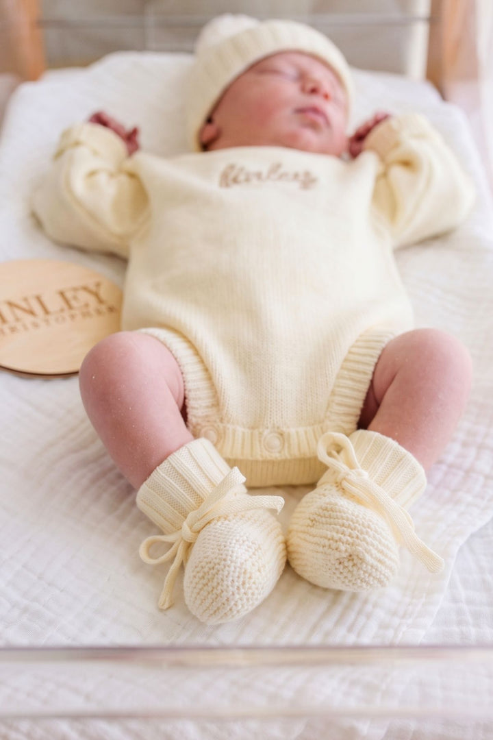 A sleeping newborn baby wearing a cream-colored knit onesie with 'Anley' embroidered on the chest, a matching knit hat, and booties, lying on a white textured blanket. A wooden brand tag for 'Anley Christchurch' is visible on the left.