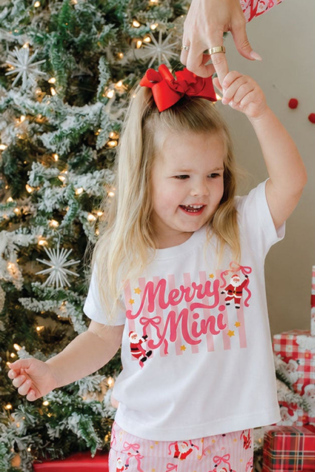 Girl wearing the Caden Lane Merry Mini Kids' Graphic Tee with Jolly Ribbons Christmas bamboo flare pants, celebrating the holidays beside a decorated Christmas tree.