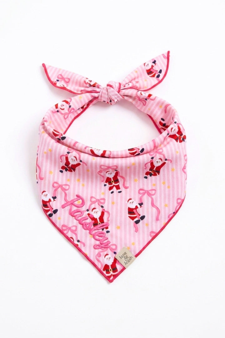 A pink and white striped dog bandana from Lane & Leash tied in a knot, featuring a whimsical print of Santa Claus figures dancing while holding pink ribbon bows, scattered with small gold stars. The name 'Paisley' is embroidered in pink cursive script near the bottom point, with a small woven Lane & Leash label tag