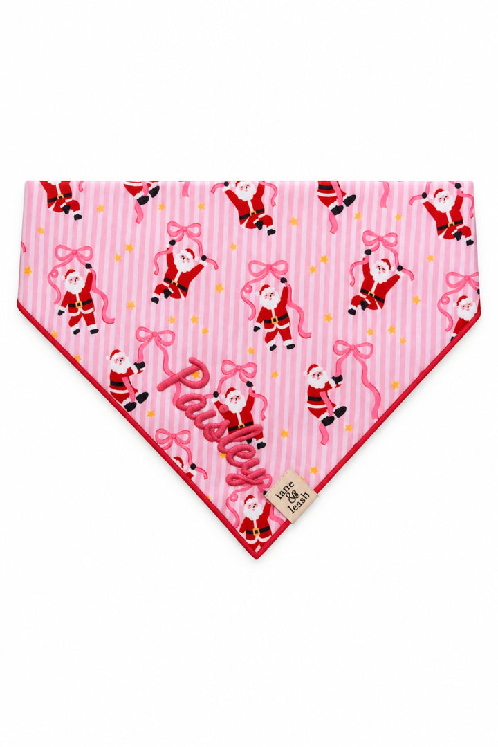 Lane & Leash personalized dog bandana in pink stripe Jolly Ribbons print with dancing Santa Claus figures and bows, tied bandana-style, embroidered with the name 'Paisley' in pink