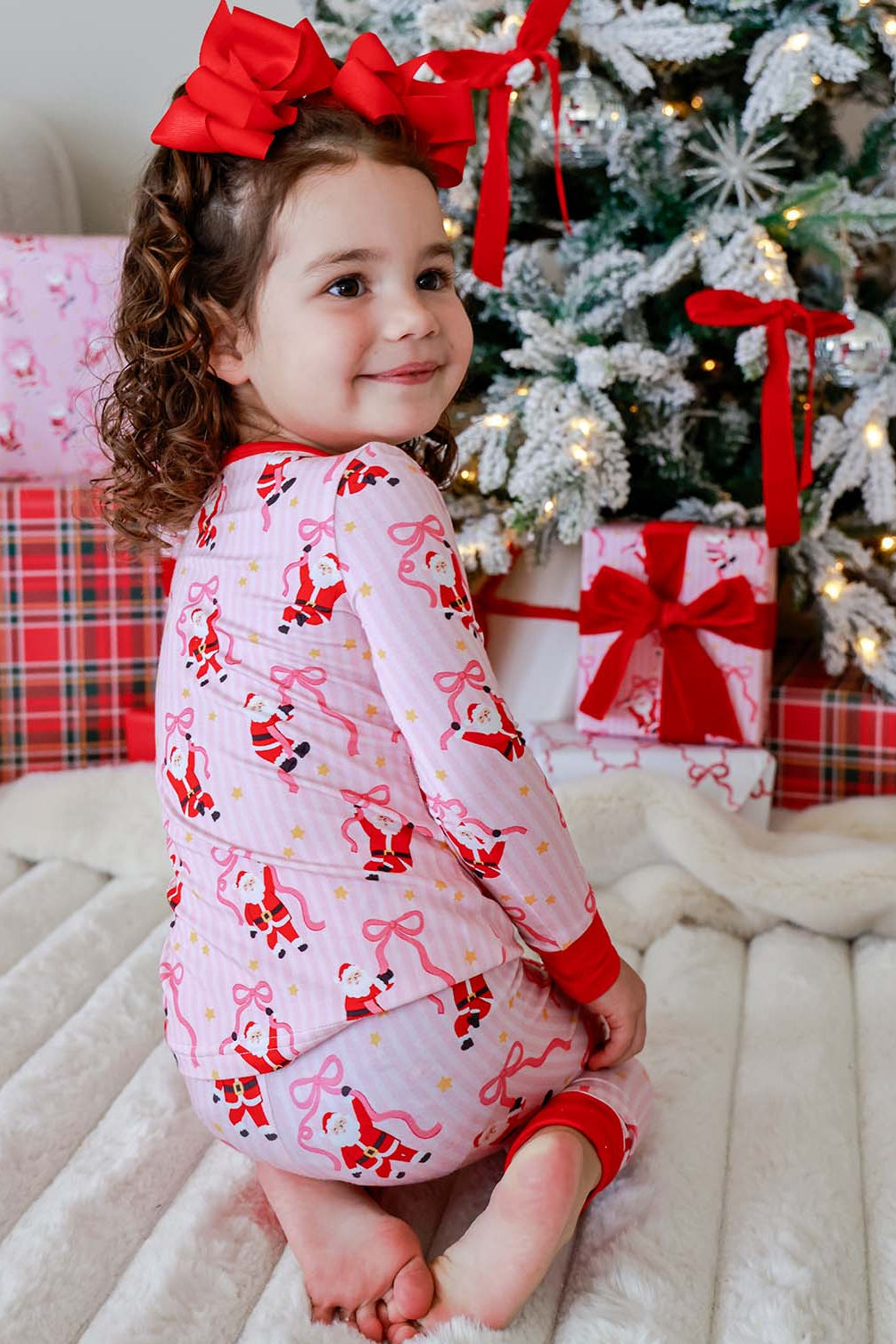 Girl wearing the Caden Lane Jolly Ribbons Christmas Bamboo Two-Piece Pajama Set beside a decorated Christmas tree, featuring festive Santa and ribbon bow print.