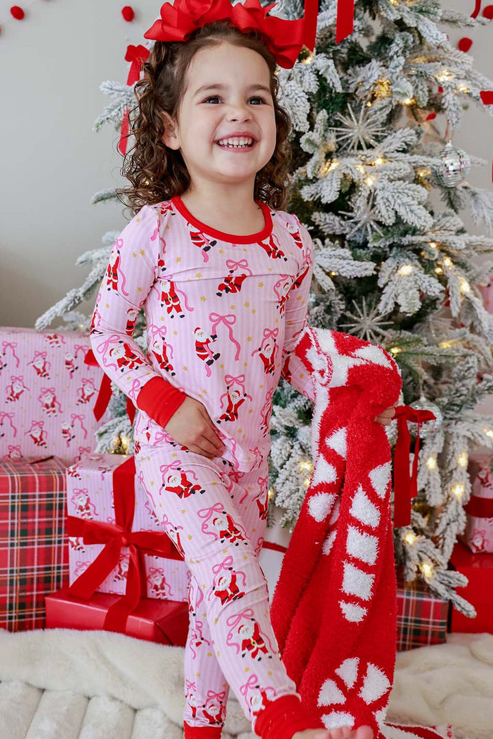 Girl wearing the Caden Lane Jolly Ribbons Christmas Bamboo Two-Piece Pajama Set, carrying a festive candy cane blanket beside a decorated Christmas tree.