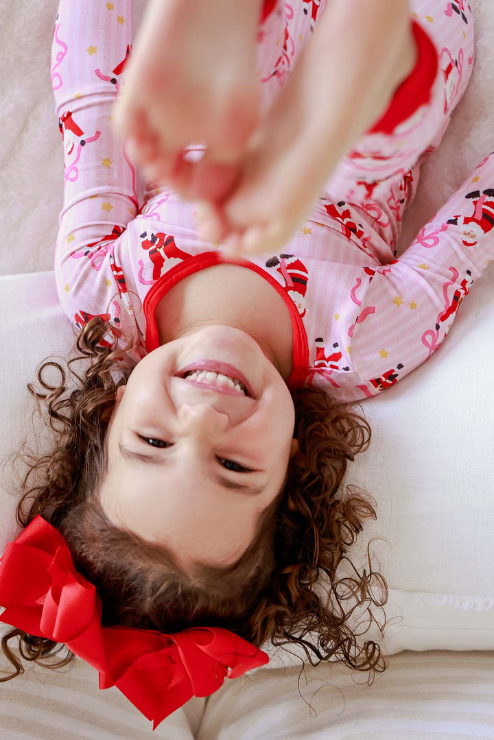 Girl wearing the Caden Lane Jolly Ribbons Christmas Bamboo Two-Piece Pajama Set with festive Santa and ribbon bow print, smiling during holiday playtime.