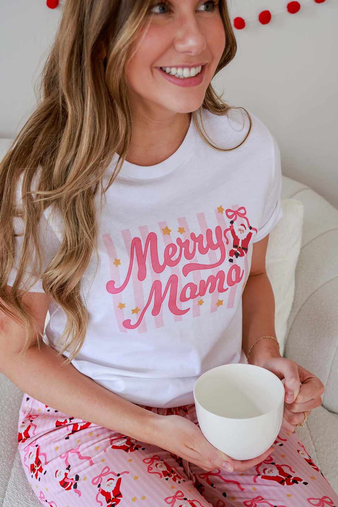 Close-up of the Caden Lane Women's Merry Mama Graphic Tee paired with Holly Jolly Christmas pajama pants, featuring a festive Santa and pink ribbon bow graphic.