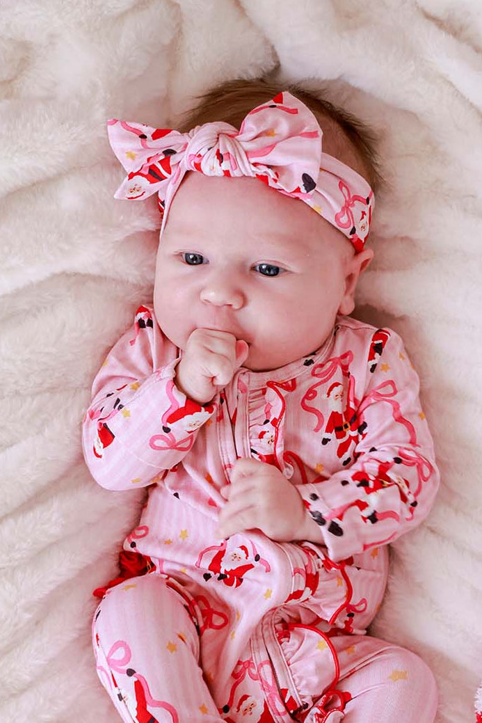 Baby wearing the Caden Lane Jolly Ribbons Christmas Knit Large Bow Headwrap with a matching Bamboo Convertible Zip Romper featuring festive Santas and ribbon bows.
