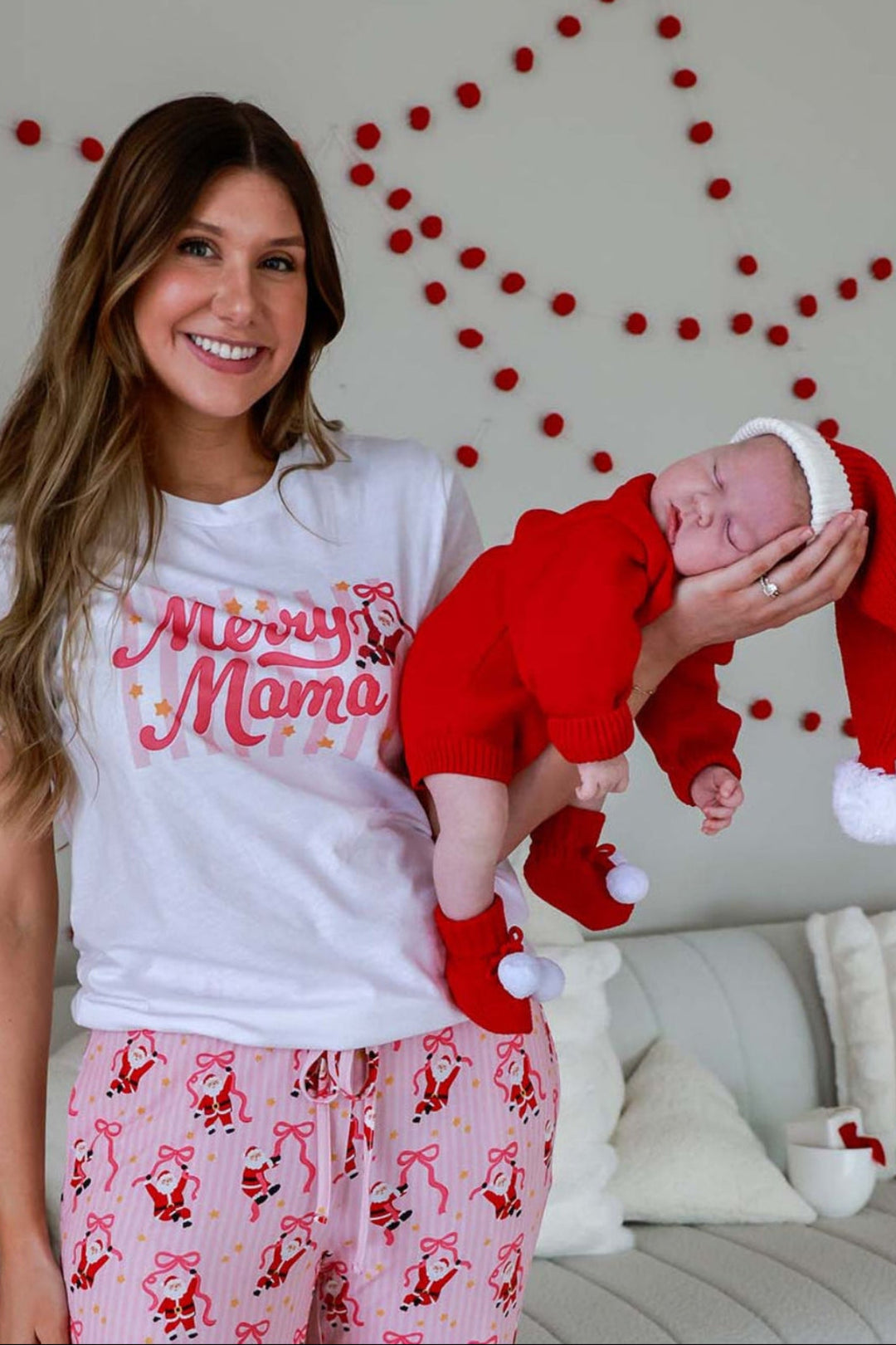 Mom wearing the Caden Lane Merry Mama Graphic Tee and matching Holly Jolly Christmas pajama pants while holding her newborn in a festive Santa outfit.