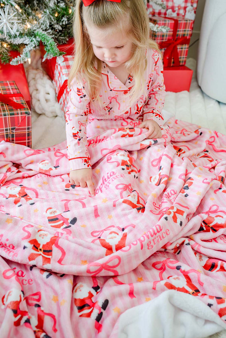 A young girl sits on the floor wrapped in a pink Christmas-themed blanket and wearing matching pajamas with Santa Claus prints, surrounded by wrapped gifts and a decorated tree.