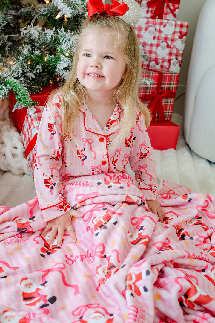 Toddler enjoying the Caden Lane Personalized Name Christmas Blanket in the Jolly Ribbons print while wearing matching bamboo Christmas pajamas, featuring personalized holiday keepsake blanket with festive Santa and ribbon pattern.