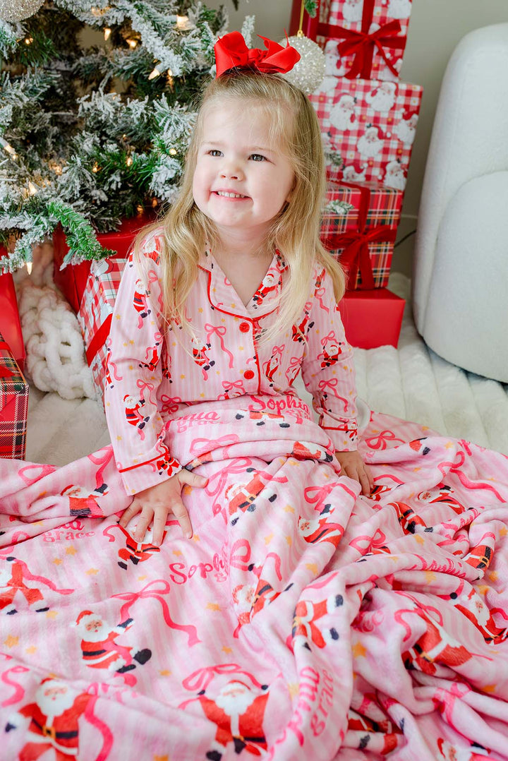 A young girl with blonde hair and a red bow sits wrapped in a pink Christmas-themed blanket, wearing matching pink pajamas. She is posed in front of a decorated Christmas tree and wrapped presents.