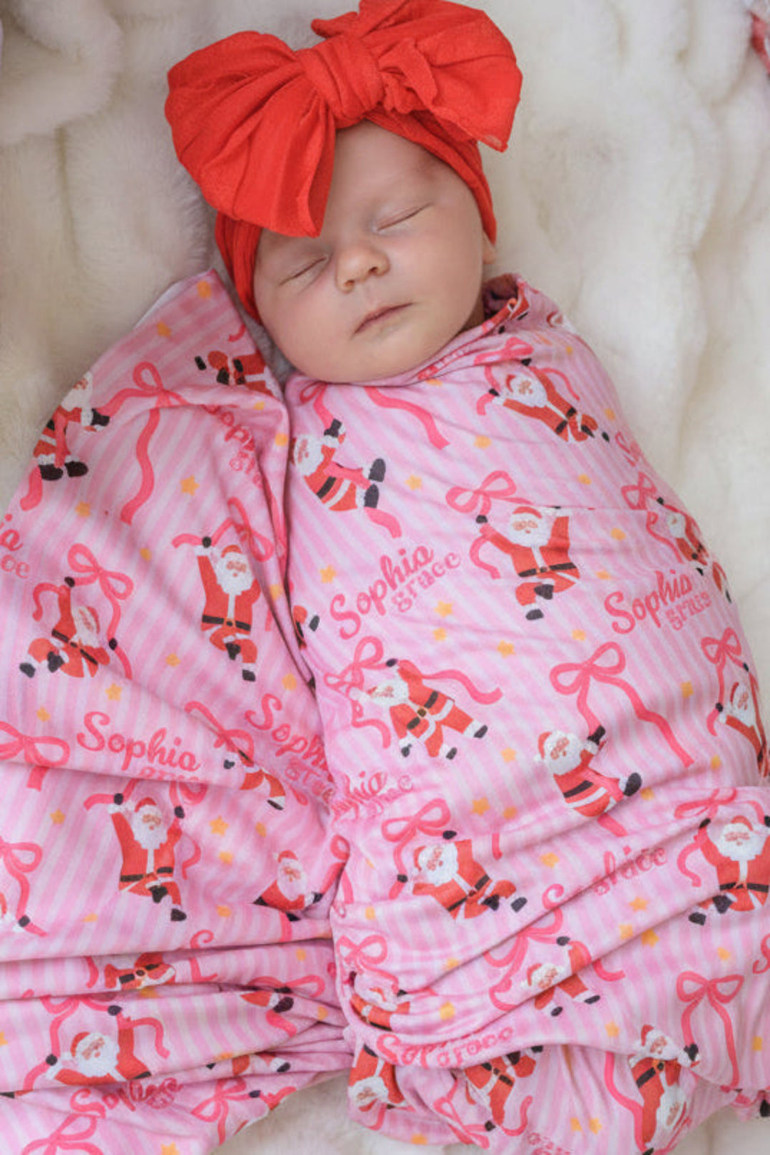 Sleeping baby wrapped in the Caden Lane Personalized Jolly Ribbons Bamboo Swaddle Blanket with custom name personalization and coordinating oversized red bow for baby's first Christmas.