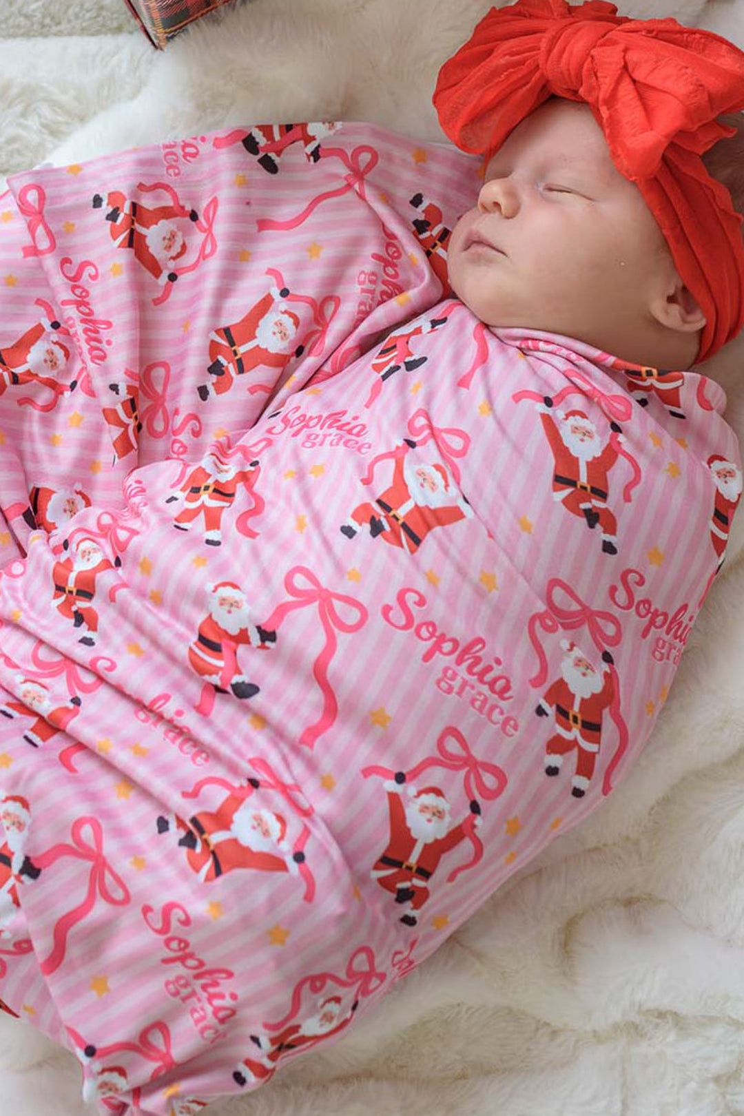 Baby wrapped in the Caden Lane Personalized Jolly Ribbons Bamboo Swaddle Blanket featuring festive Santas, ribbon bows, custom name personalization, and a coordinating oversized red bow.
