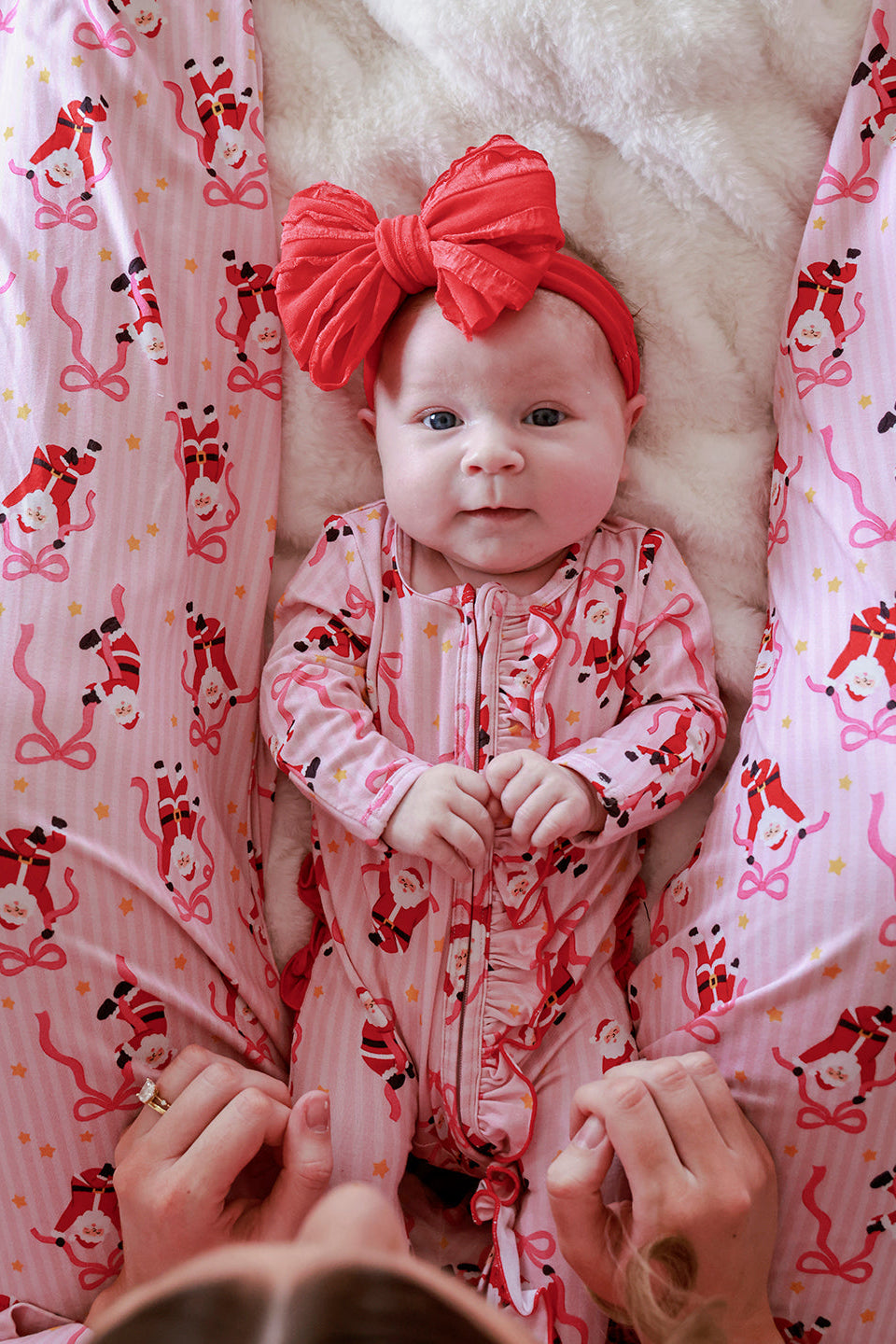 Baby wearing the Caden Lane Jolly Ribbons Christmas Zipper Ruffle Footie with a coordinating oversized red bow, featuring festive Santas and ribbon bows.