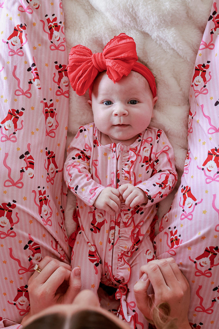 Baby wearing the Caden Lane Jolly Ribbons Christmas Zipper Ruffle Footie with a coordinating oversized red bow, featuring festive Santas and ribbon bows.