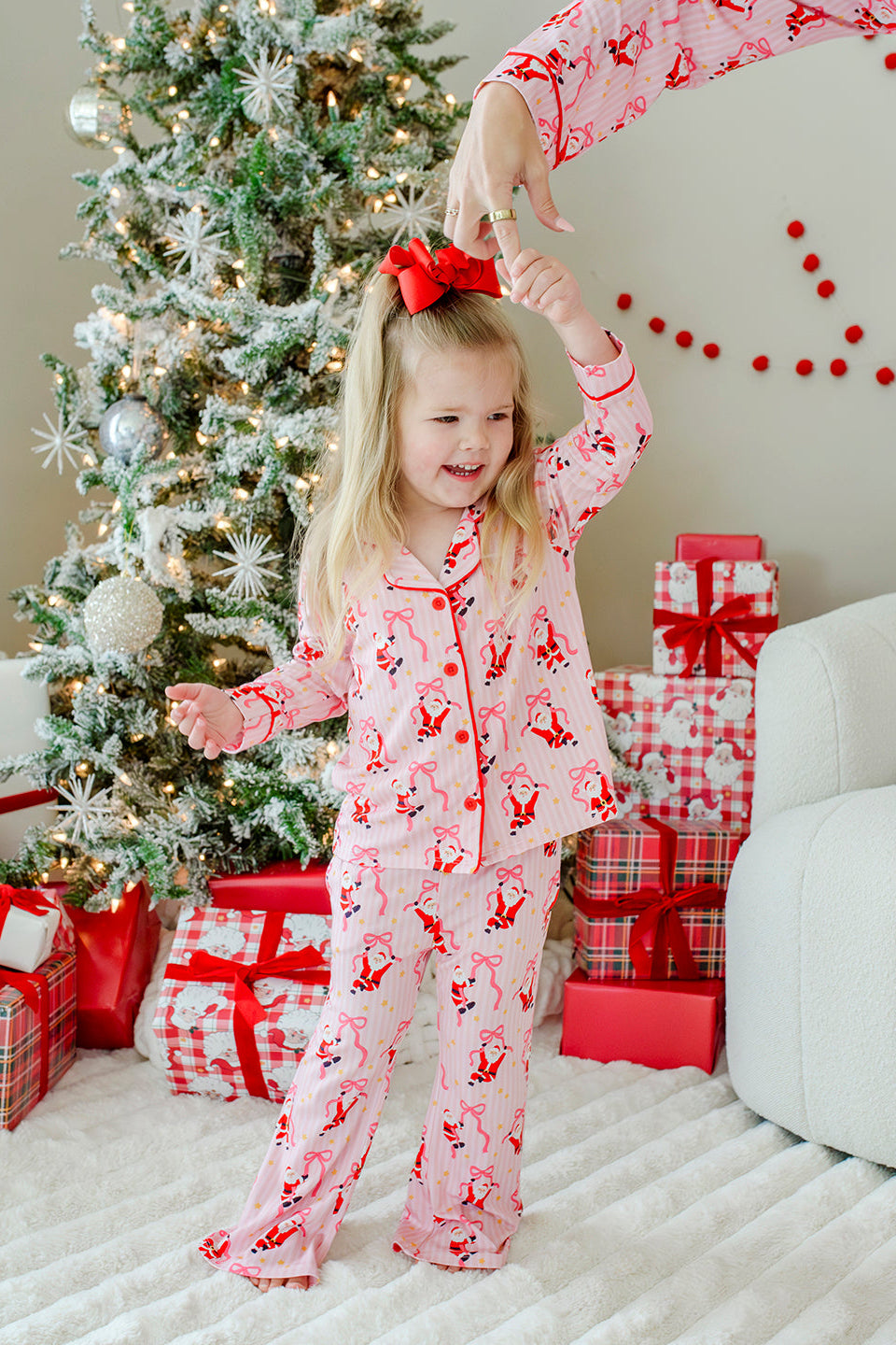 Girl wearing the Caden Lane Jolly Ribbons Girls' Christmas Two-Piece Flare Lounge Set featuring festive Santas and ribbon bows while twirling beside a decorated Christmas tree.