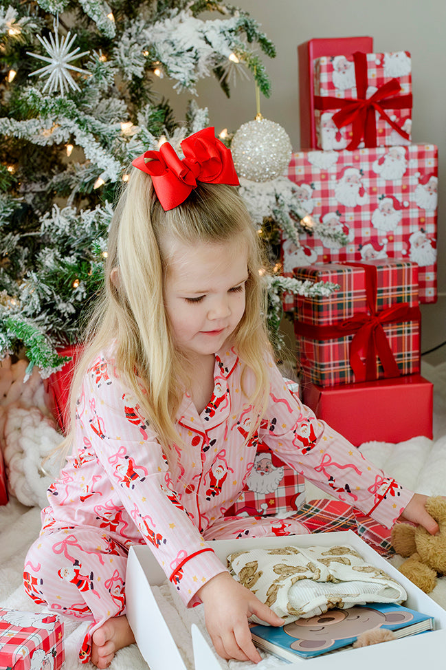 Girl wearing the Caden Lane Jolly Ribbons Christmas Bamboo Two-Piece Pajama Set opening Christmas gifts, featuring festive Santas, ribbon bows, and buttery-soft bamboo fabric.