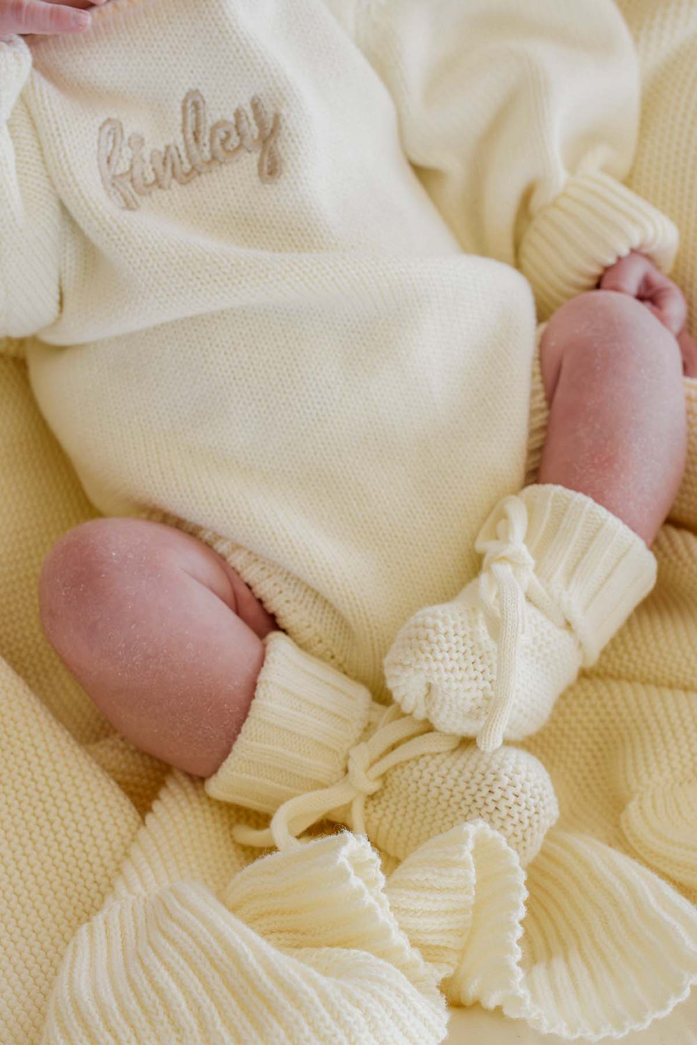 Close-up of Caden Lane ivory knit bubble romper with "Finley" name embroidery and matching tie-front pom-pom booties