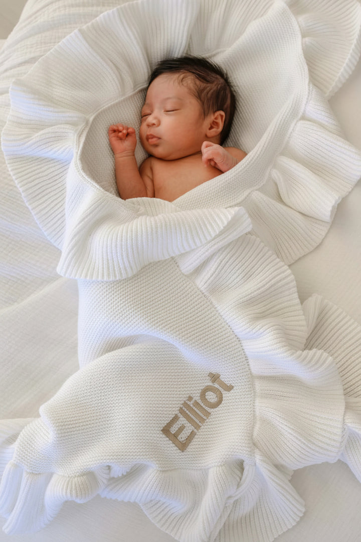 A newborn baby wrapped in a white textured blanket with ruffled edges, lying on a white surface. The blanket is personalized with the name 'Elliot' in a light brown embroidered font.