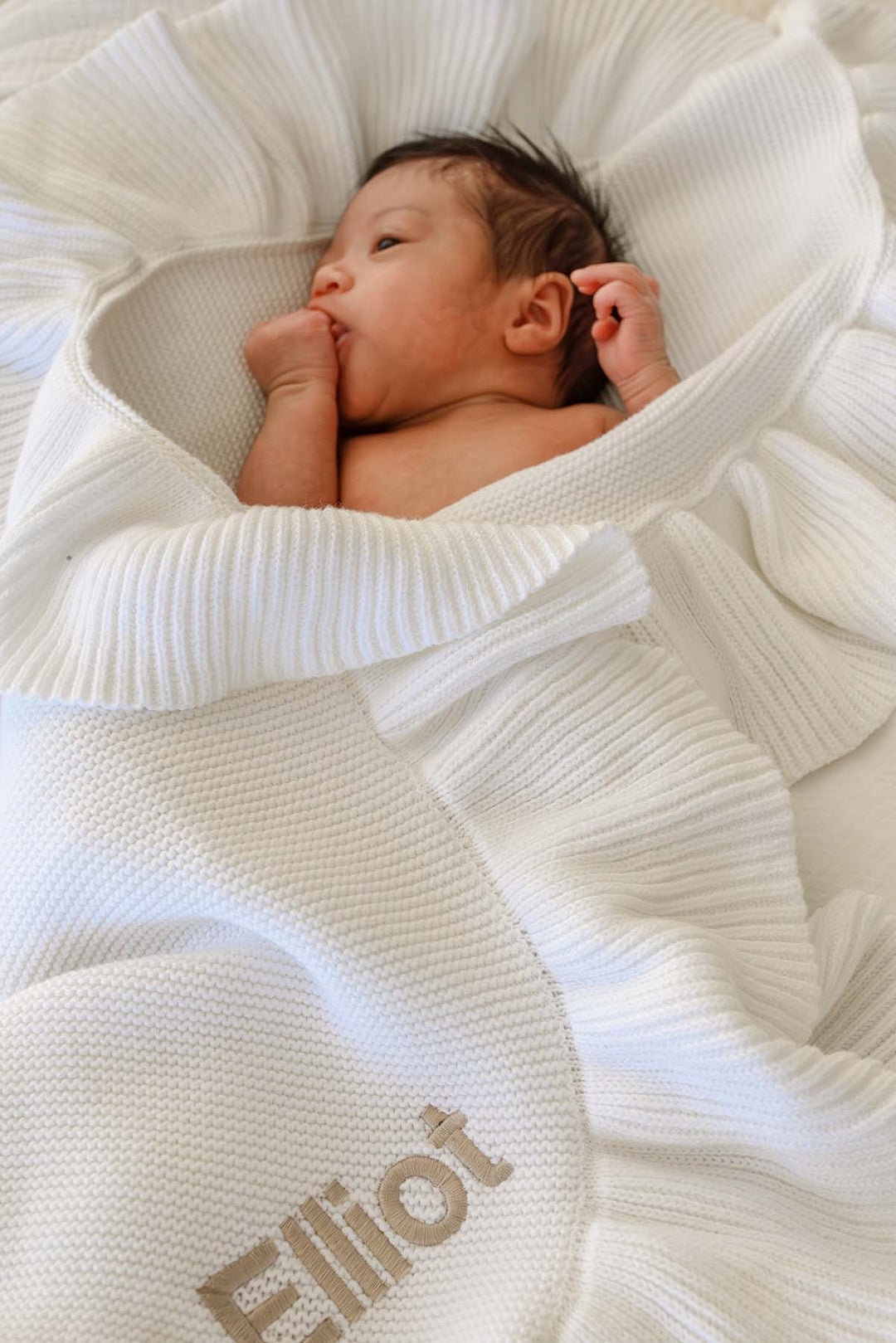 A newborn baby wrapped in a white ribbed knit blanket with the name 'Elliot' embroidered in beige, lying on a white textured blanket.