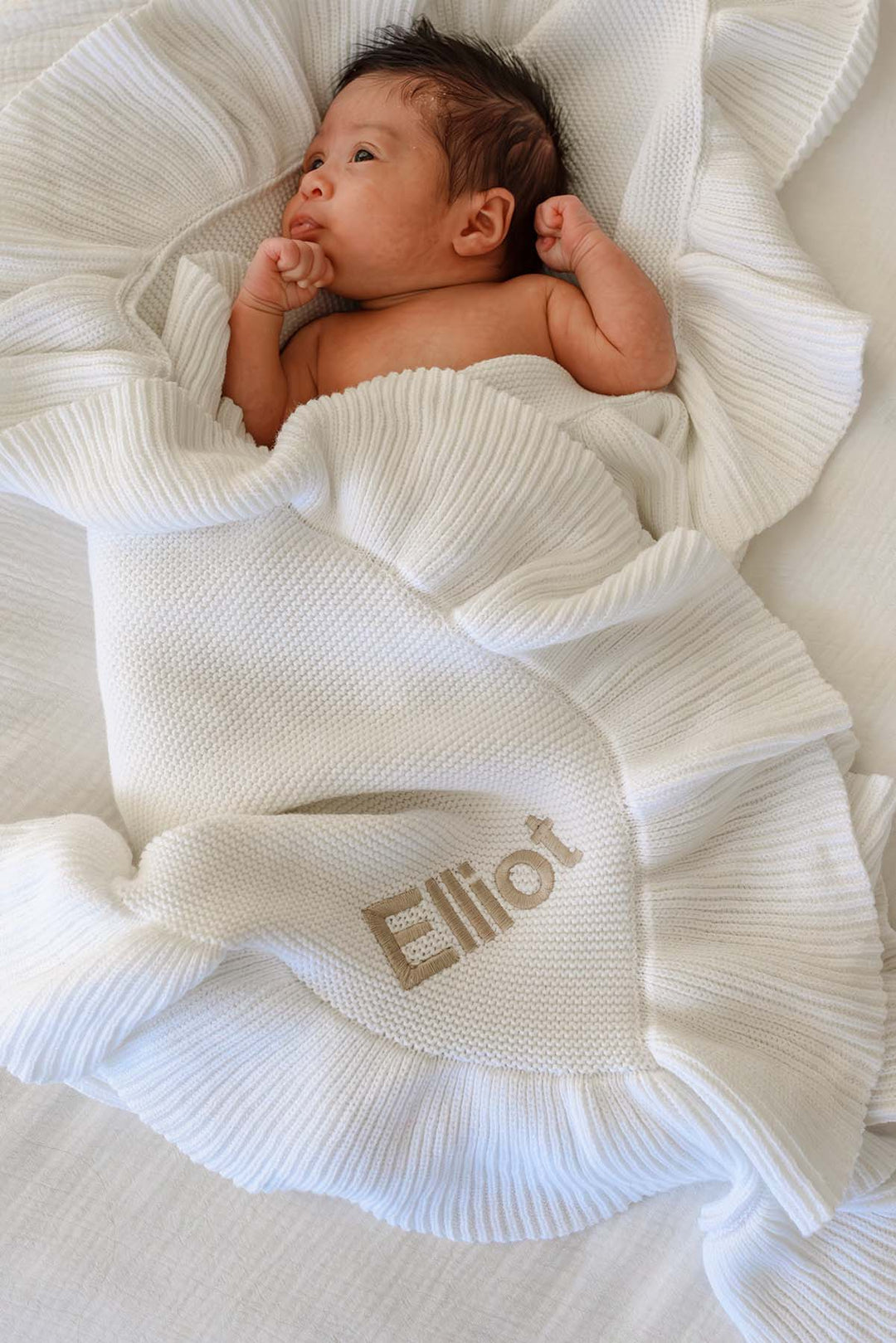 A newborn baby wrapped in a white textured knit blanket with the name 'Elliot' embroidered in gold, lying on a white ruffled blanket.