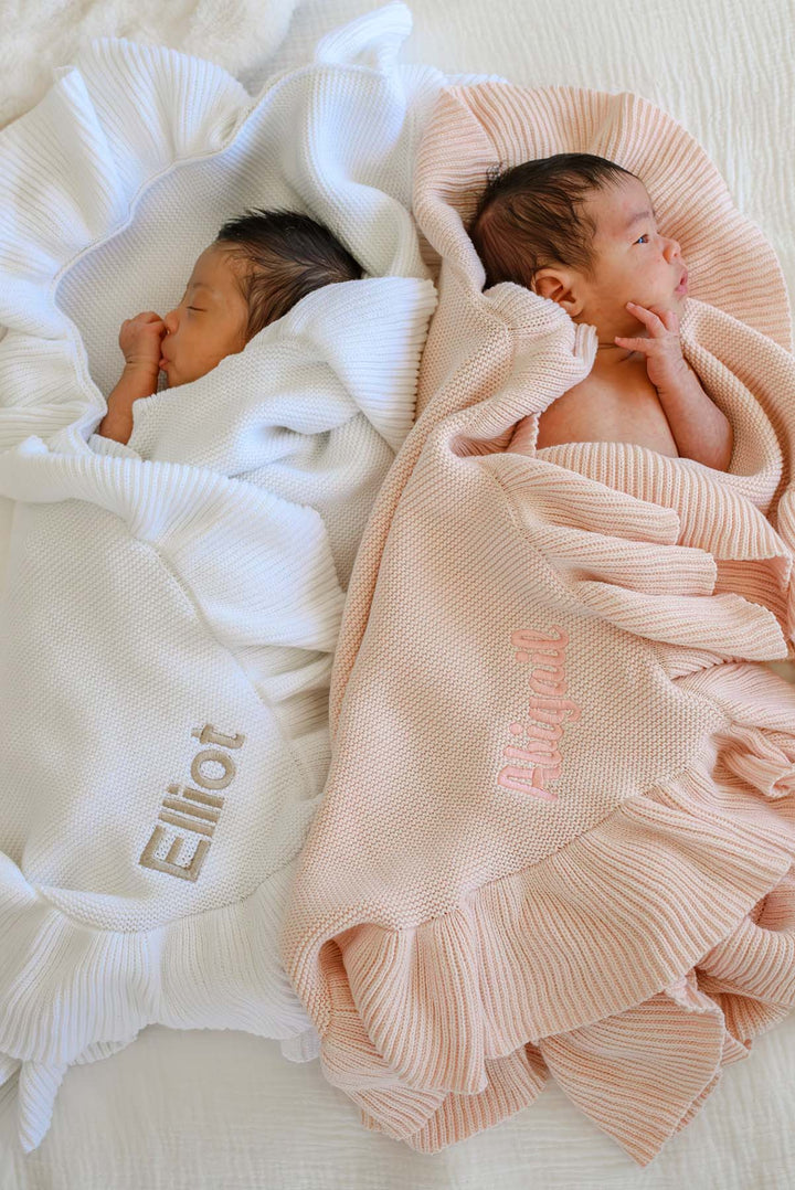 Two newborn babies sleeping on a white textured blanket, each wrapped in a ribbed knit swaddle. The left swaddle is white with the name 'Elliot' embroidered in gray, and the right swaddle is pale pink with the name 'Alina' embroidered in pink.