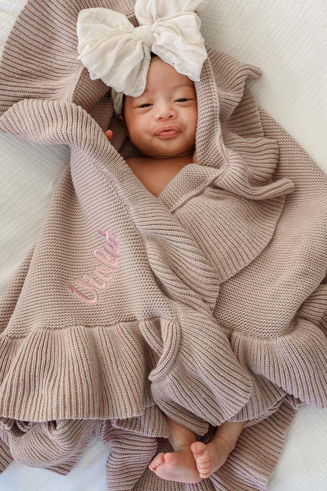 A newborn baby wrapped in a beige ribbed knit hooded blanket with ruffled edges, featuring a large cream bow on the hood and pink embroidered text on the front, lying on a white blanket.