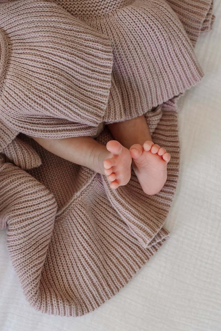 A close-up of a baby's bare feet peeking out from under a soft, ribbed knit blanket in a dusty rose or mauve color, laid on a white textured blanket.
