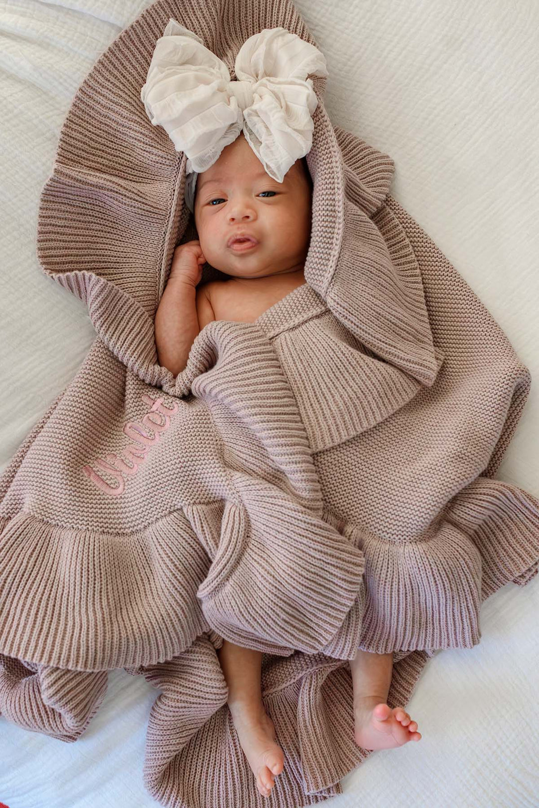 A baby wrapped in a chunky ribbed knit blanket with a large matching bow on the hood, lying on a white textured blanket. The blanket has pink embroidered text reading 'braves'.