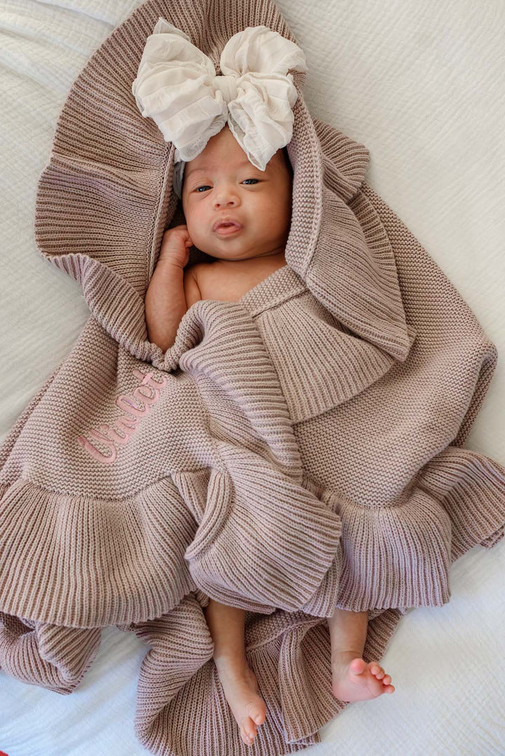 A baby wrapped in a chunky ribbed knit blanket with a large matching bow on the hood, lying on a white textured blanket. The blanket has pink embroidered text reading 'braves'.