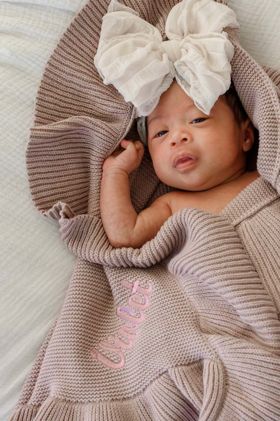 A baby wrapped in a soft ribbed knit blanket with a large white bow on the head, lying on a light textured surface. The blanket has pink embroidered text reading 'Love, Love, Love'.
