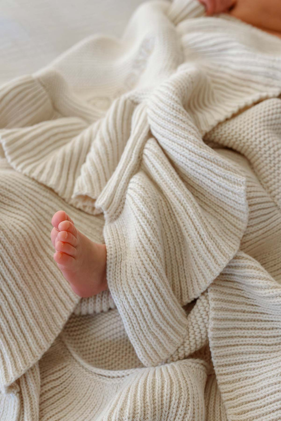 A close-up of a soft cream-colored knit blanket with ribbed and waffle textures, with a baby's bare foot peeking out from underneath.