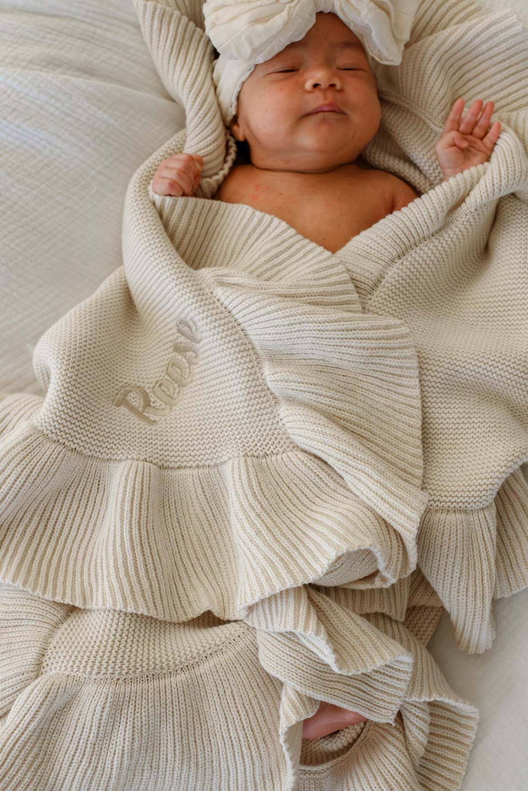 A sleeping newborn baby wrapped in a cream-colored ribbed knit blanket with a matching turban hat, on a soft neutral background.