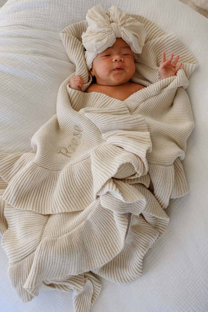 A newborn baby wrapped in a cream-colored ribbed knit blanket with a matching knotted turban hat, lying on a white textured blanket.