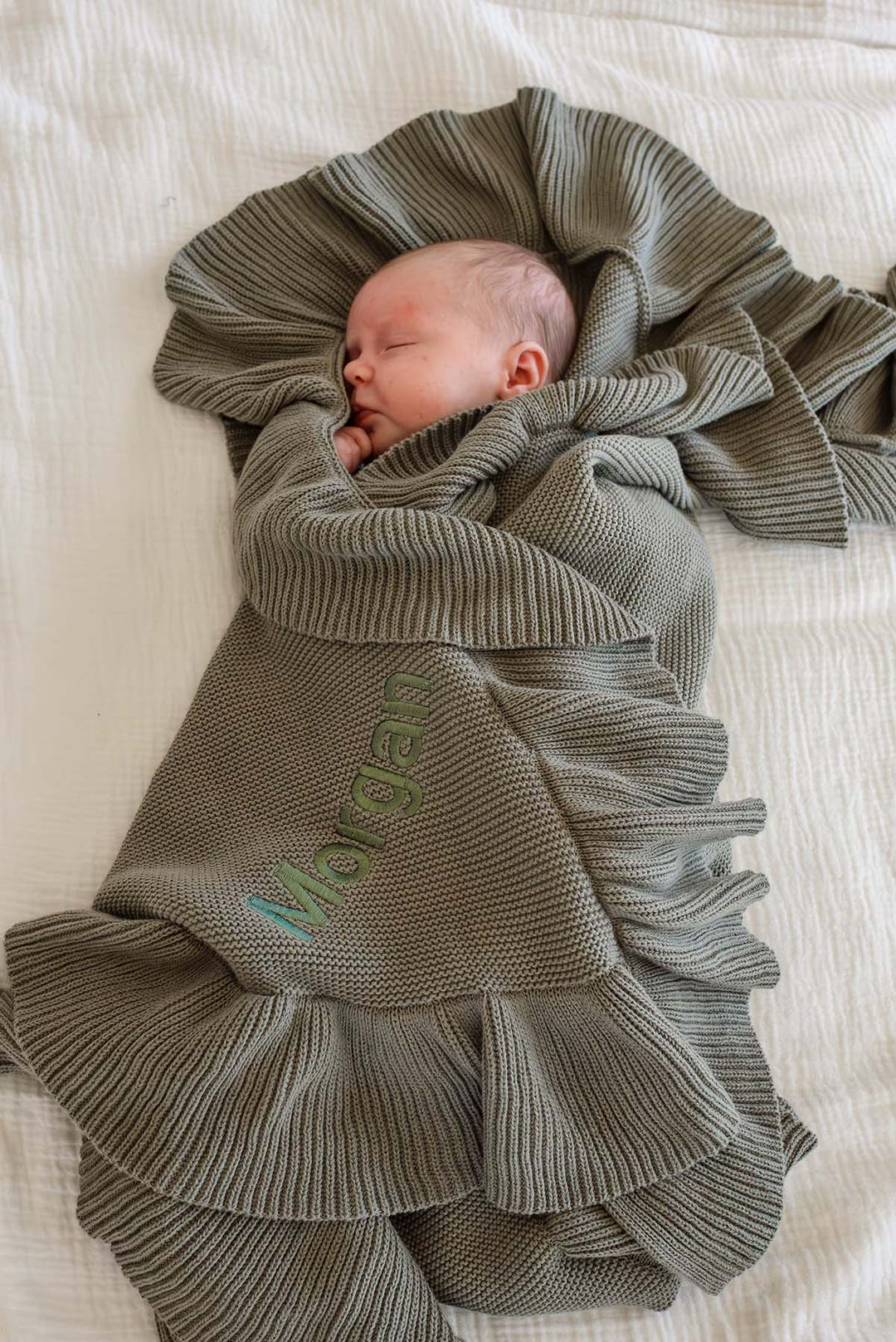 A sleeping newborn baby is swaddled in a ribbed knit blanket with ruffled edges, laid on a white textured bed. The blanket is personalized with the name 'Morgan' in green embroidery.